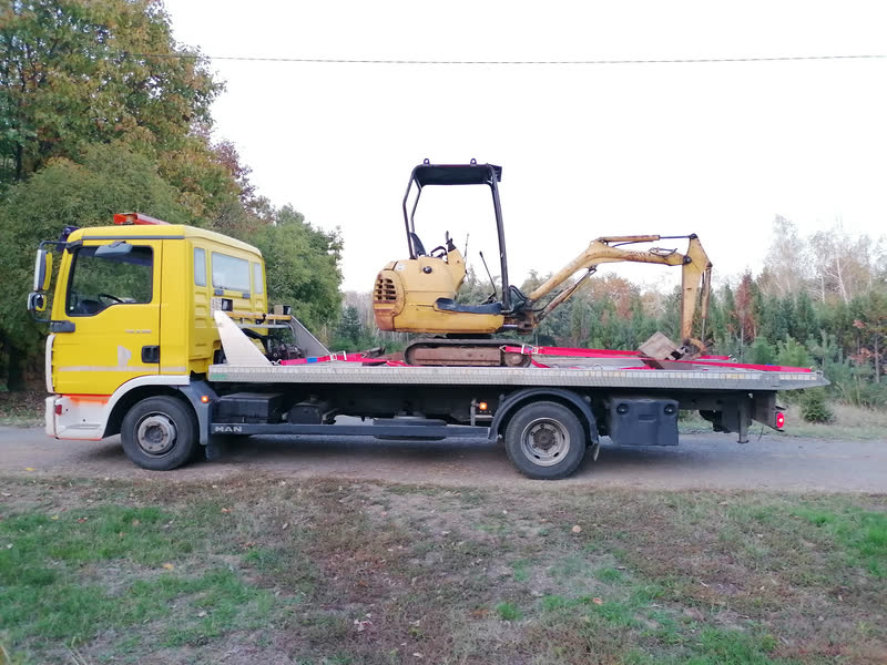 Žuti MAN šleper iz profila, sa mini bagerom vezanim trakama na platformi.