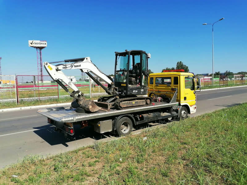 Beli bager Bobcat na platformi žutog MAN šlepera, parkiranog uz put, po sunčanom danu.