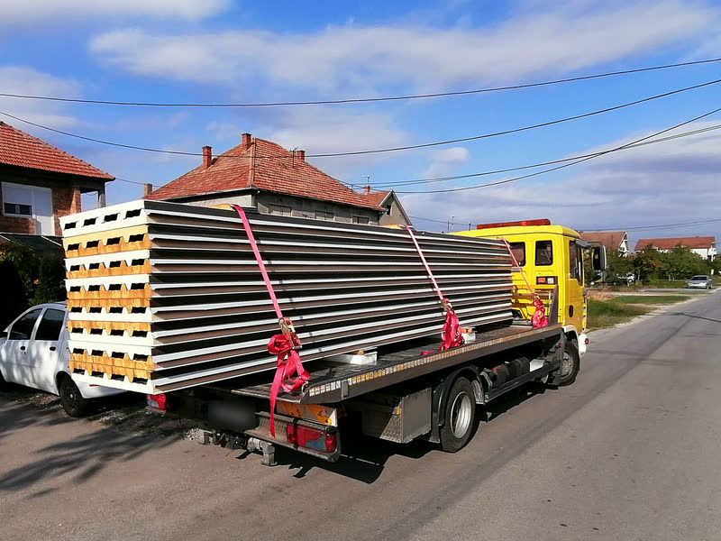 Paleta sendvič panela vezana crvenim trakama na platformi šlepera, u ulici sa kućama.